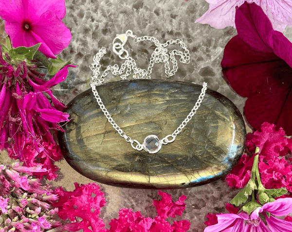 The Amethyst Tiny Drop on a custom 16" vintage sterling silver chain by Amazing Earth rests on an iridescent stone, surrounded by vibrant pink and red flowers—a stunning crystal jewelry piece.