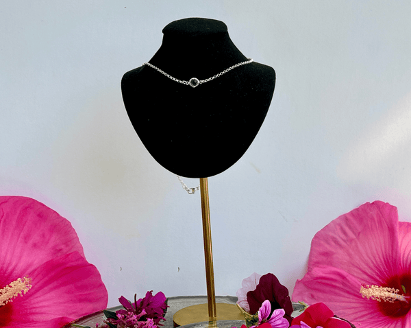 The Amethyst Tiny Drop on Custom 16 in Vintage Sterling Silver Chain by Amazing Earth is displayed on a black velvet stand with a gold base, accented by large pink and red flowers at the stand’s base against a white background.