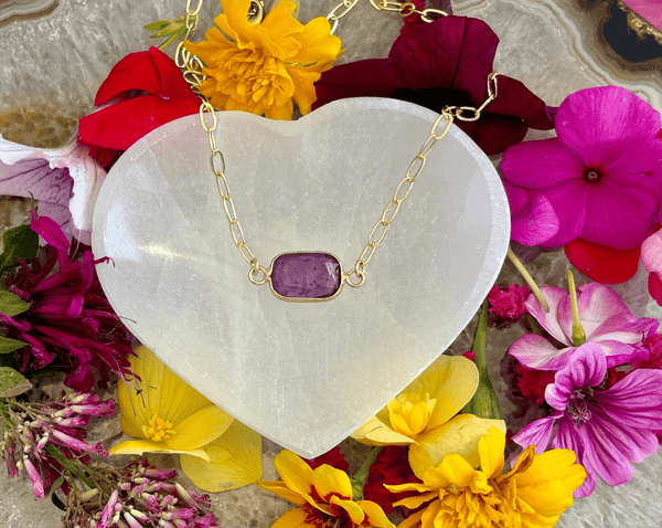 The Amazing Earth Amethyst on 16 in 14KGF Vintage Paper Clip Chain necklace is displayed on a white heart-shaped stone, surrounded by vibrant yellow, pink, red, and purple flowers—a stunning crystal jewelry piece.