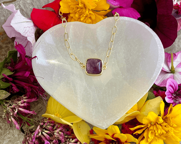 The Amethyst Square on 16 in 14KGF Vintage Paper Clip Chain by Amazing Earth is displayed on a heart-shaped white stone, surrounded by pink, yellow, and red flowers—a striking crystal jewelry piece.