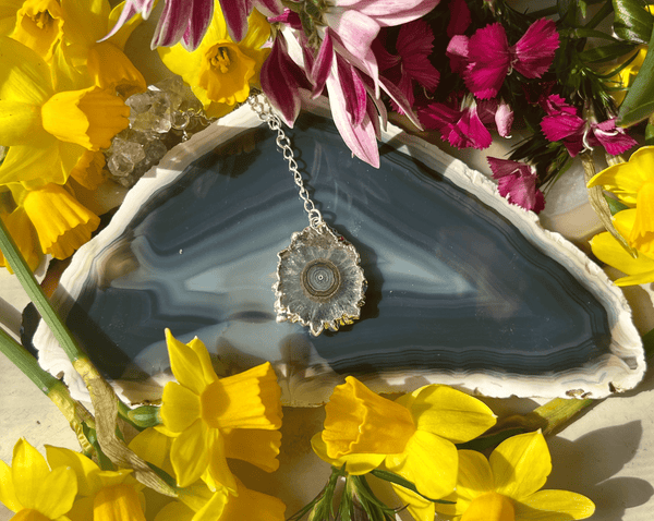 The Amethyst Stalactite Dropping from Sterling Chain by Amazing Earth features a crystal-like pendant on polished amethyst, surrounded by yellow daffodils and pink flowers—a stunning handmade crystal jewelry piece.