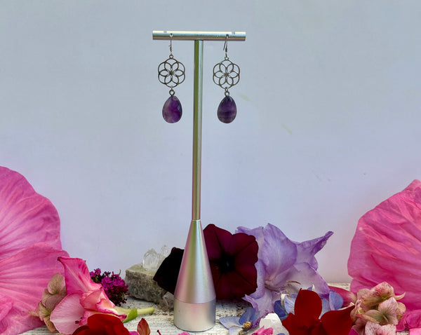 Flower Earrings with Chevron Amethyst Teardrops by Amazing Earth feature geometric sterling silver backs and purple stones, displayed on a silver stand with vibrant flowers and petals in the background.