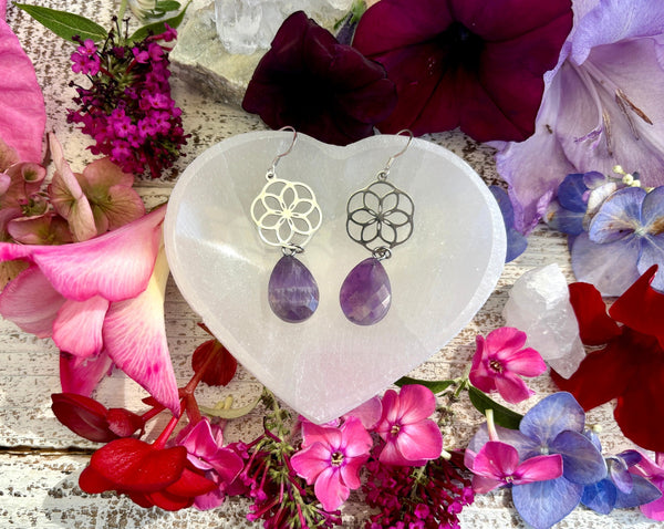 Amazing Earth’s Flower Earrings with Chevron Amethyst Teardrops on sterling silver backs are displayed on a heart-shaped stone among colorful flowers and petals atop a rustic white wooden surface—a striking crystal jewelry piece.
