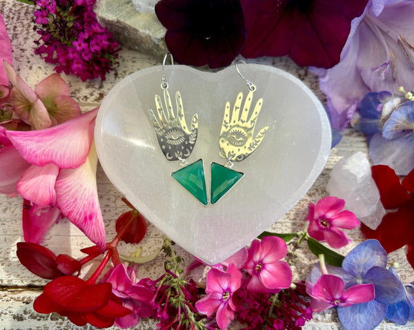 The Hand and Protective Eye Earrings by Amazing Earth feature sterling silver with green onyx triangles and are shown on a heart-shaped white dish, surrounded by vibrant flowers and crystal jewelry on rustic wood.