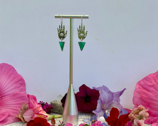 A pair of Amazing Earth's Hand and Protective Eye Earrings with sterling silver green onyx triangles rest on a stand, surrounded by pink, red, and purple flowers against a plain white background.