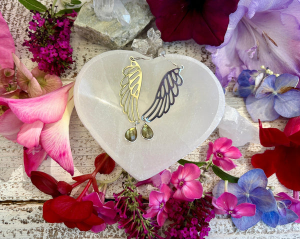 Amazing Earth Angle Wing Earrings feature sterling silver labradorite teardrops and secure sterling silver backs, displayed on a heart-shaped stone among colorful flowers and crystals atop rustic wood.