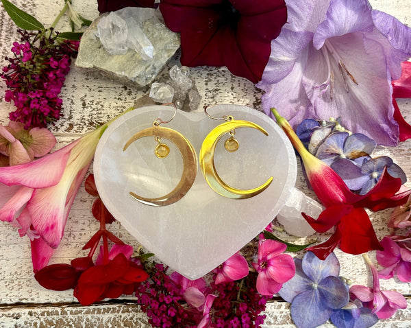 Amazing Earth’s Half Moon Earrings with Gold Citrine Dangles on 14KG backs are displayed on a heart-shaped white stone, surrounded by colorful flowers, citrine drops, and crystals atop a rustic white wooden surface.