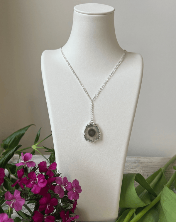 The Amazing Earth Amethyst Stalactite Dropping from Sterling Chain is showcased on a white jewelry bust, featuring a circular pendant with a dark amethyst center and surrounded by green leaves and purple flowers.