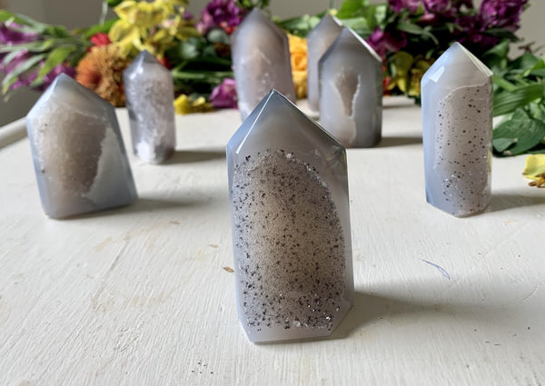 Amazing Earth's Agate Towers with Druzy Windows, featuring polished gray and white crystal points, are displayed on a white surface with softly blurred colorful flowers in the background.
