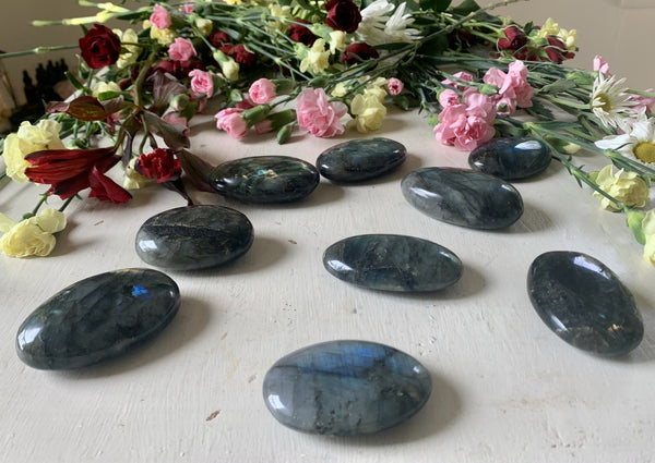 Amazing Earth Labradorite Palm Stones, smooth and oval, are displayed on a white surface with pink, red, and yellow flowers behind them. These stunning stones reflect blue and green hues and are perfect for intuition work.