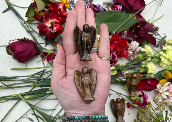 A hand holds two Amazing Earth angel figurines, including a Tigers Eye Crystal Angel for protection. Another angel rests on the palm, with colorful flowers and greenery scattered on a white background.