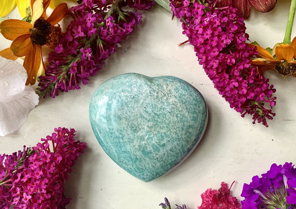 The Amazing Earth Amazonite Crystal Heart, a soothing symbol of wealth manifestation, is displayed on a light surface surrounded by vibrant purple, orange, and pink flowers.