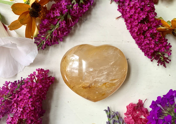 The Amazing Earth Golden Healer Crystal Heart rests on a white surface among vivid purple flowers and colorful petals, creating an inviting display that symbolizes energy healing and self-care.