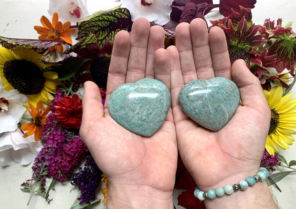 Two hands hold polished, heart-shaped stones—including the Amazing Earth Amazonite Crystal Heart—above a vibrant floral display. One wrist features a teal and white beaded bracelet, accenting the crystal’s soothing energy.