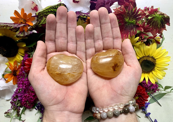 Two hands hold polished Amazing Earth Golden Healer Crystal Hearts, surrounded by colorful flowers and greenery. A beaded bracelet on the left wrist highlights the Golden Healer’s energy healing properties.