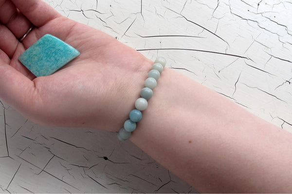 A hand wearing a light blue amazonite energy bracelet holds a large blue-green rectangular stone. The background is a white surface with black cracks, highlighting this calming crystal often associated with the heart chakra.