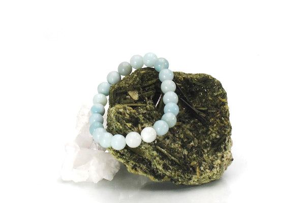A light blue amazonite energy bracelet, known as a calming crystal for the heart chakra, rests on a large, rough greenish-brown mineral rock with a smaller white crystal beside it, all set against a white background.