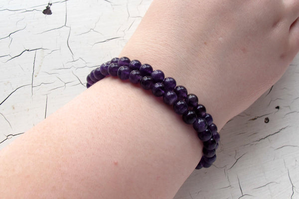A hand wears two stacked Amazing Earth Amethyst Energy Bracelets, featuring small, shiny, round purple stones, on a white cracked surface. Amethyst is known as a protective stone and energy bracelet.