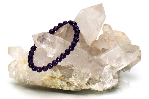 The Amazing Earth Amethyst Energy Bracelet, featuring purple beads, is displayed on a clear quartz crystal cluster against a white background, highlighting the bracelet's natural allure.