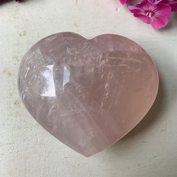 The Rose Quartz Crystal Heart by Amazing Earth rests on a light surface, with a few pink flowers in the upper right corner.