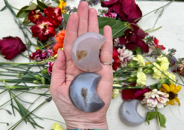 A hand holds two Amazing Earth Agate Crystal Moon with Druzy Window pieces above a table sprinkled with red, yellow, and purple flowers, while another druzy agate moon for anxiety relief rests nearby.