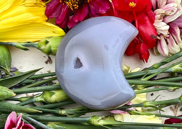 The Amazing Earth Agate Crystal Moon with Druzy Window rests on green flower stems amid yellow, red, and purple blooms, making it a charming accent piece believed to help with anxiety relief.
