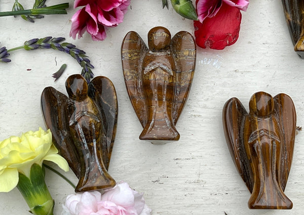 Three Amazing Earth Tigers Eye Crystal Angels are displayed on a light surface, encircled by vibrant red, pink, yellow, and purple flowers—ideal for crystal protection and peaceful decor.