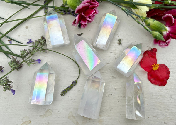 Amazing Earth Angel Aura Quartz Polished Points with rainbow reflections are displayed on a white surface, surrounded by vibrant flowers and green stems for a colorful, high-vibration crystal arrangement.