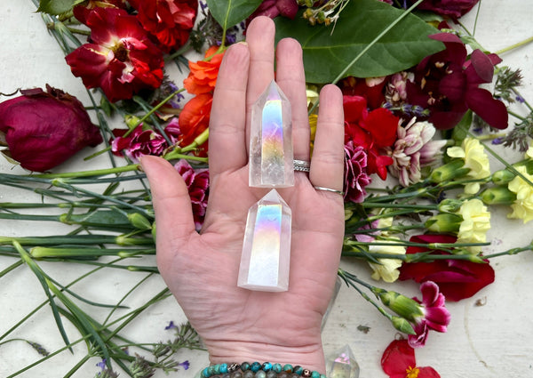 A hand adorned with silver rings and turquoise beaded bracelets holds two Amazing Earth Angel Aura Quartz Polished Points above vibrant red, purple, and pink flowers with green leaves. Rainbow reflections shimmer on the crystals.