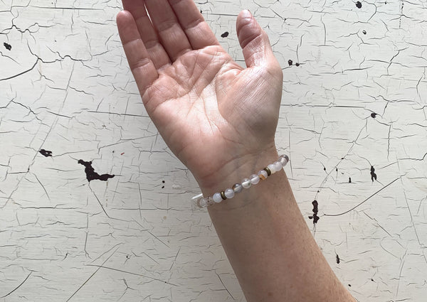A hand with the palm up wears Amazing Earth's Agate Energy Bracelet, featuring round, light stones for protection and energy cleansing, set against a white, cracked textured background.